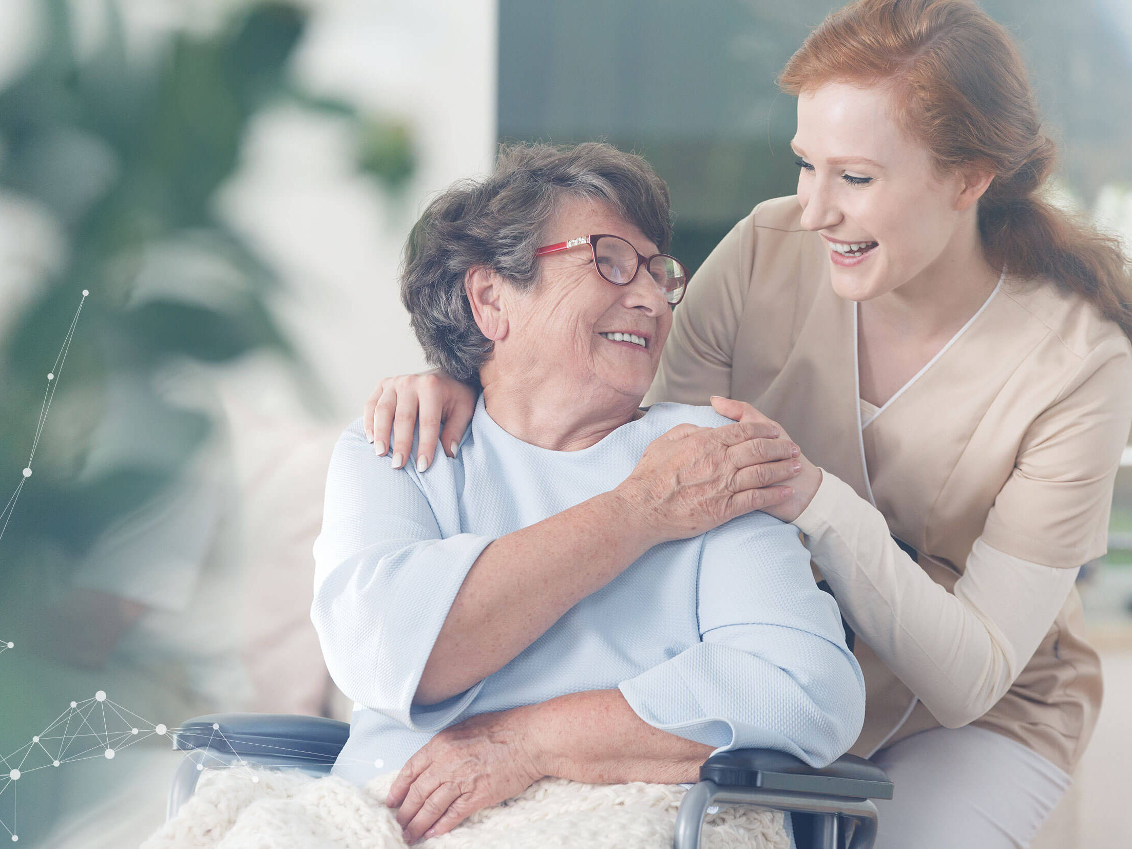 A female health-care worker standing behind a female elderly patient who is in a wheelchair. They are smiling at each other