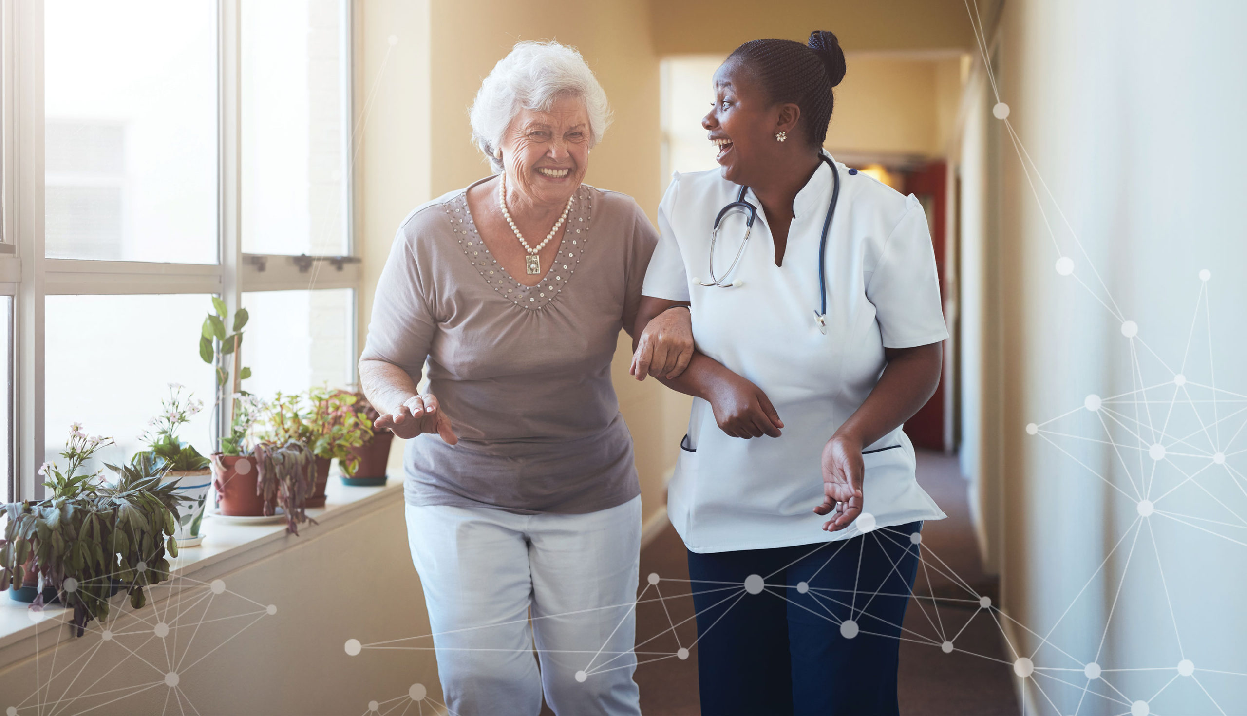 An elderly female patient walking arm in arm with a female health care worker. Both are smiling